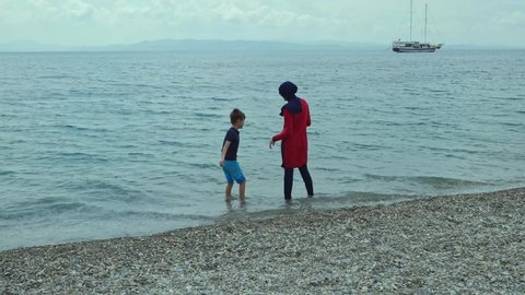A young woman in a hijab swimsuit is having fun in the sea with her little son, throwing stones into the sea. Beautiful and peaceful holiday concept. 