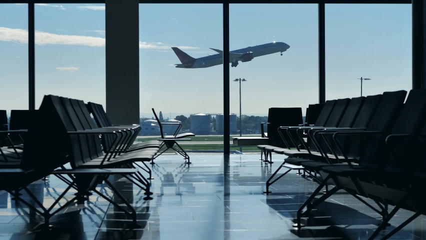 Airplane taking off seen through airport terminal window. View from inside empty waiting hall benches with no people in the lounge. Airplane departing with nobody at the gate. Royalty-Free Stock Footage #1075144439