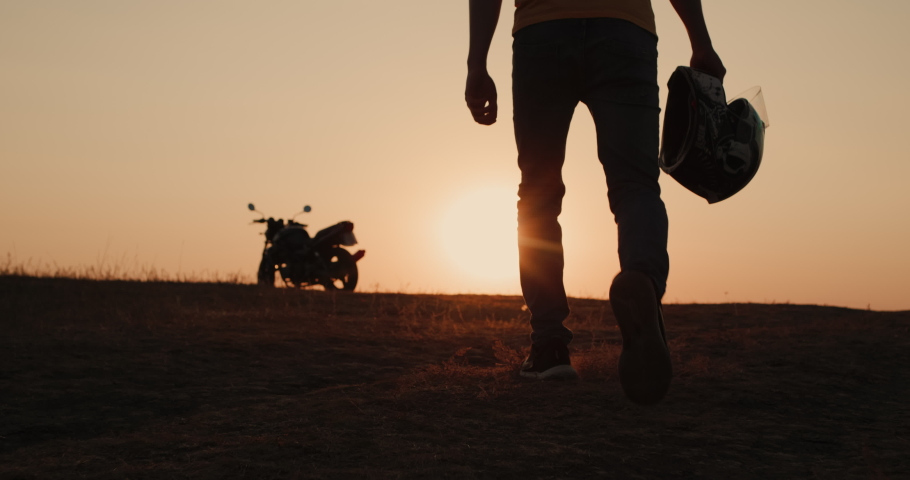 The silhouette of a motorcyclist with a helmet in his hand, goes to his motorcycle at sunset | Shutterstock HD Video #1075144958