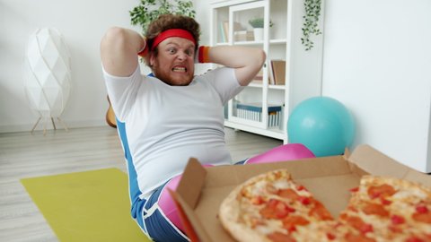 Slow motion of chubby sportsman in bright outfit doing press-ups and stretching hands to pizza training alone at home. Food and sports concept.