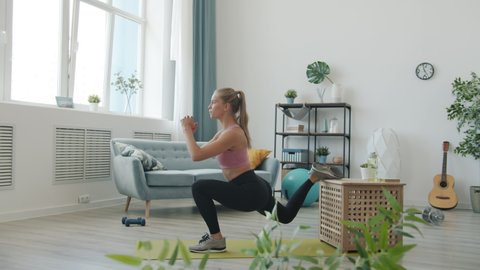 Side view of beautiful young woman in stylish sportswear doing squats standing on one leg training alone at home. Body care and fitness concept.