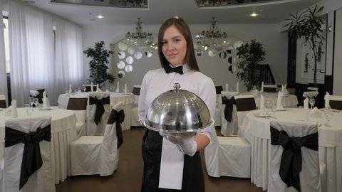 A woman waiter holds a tray with a covered dish in a restaurant.