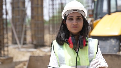 Portrait of Indian female industrial worker on construction site.Asian female industrial worker looking at camera wearing high vest and  hard hat helmet or earmuff with crossing her hands.