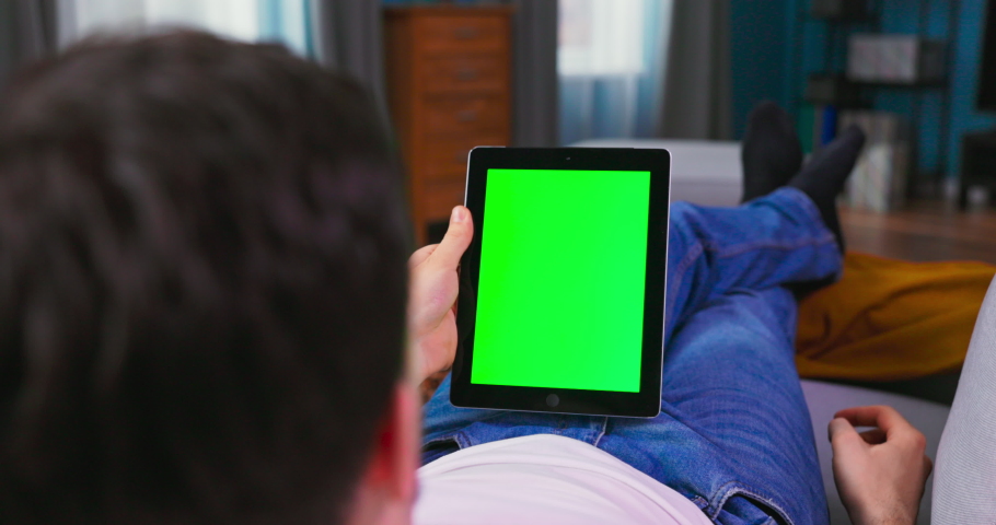Young man at Home Resting on a Couch Using with Green Mock-up Screen Tablet Computer in Horizontal Landscape Mode. Boy Using Gestures with Touchscreen Device, Browsing Internet, Watching Content.