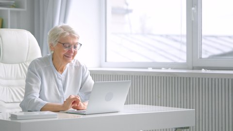 Smiling old woman secretary with grey hair types on laptop