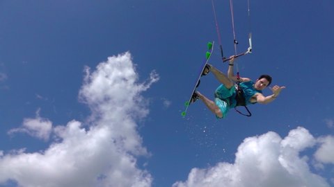 SLOW MOTION, PORTRAIT: Extreme kiteboarder jumping over the camera, showing shaka hang loose sign. Kitesurfer having fun riding the crystal clear lagoon and jumping high in the air in windy Zanzibar