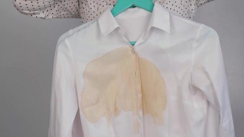 A woman compares two white shirts before and after washing. The girl is holding one blouse, clean and ironed, and the other, dirty with coffee stains
