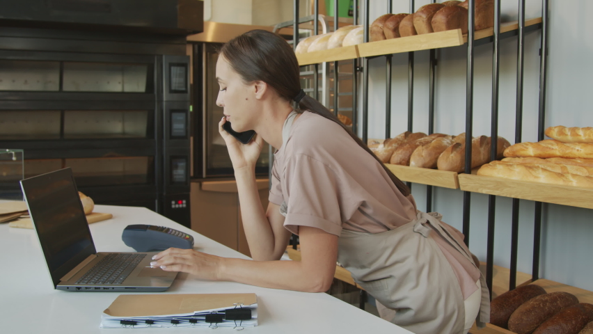 Slowmo tracking shot of cheerful young female counter attendant talking on mobile phone and using laptop while working in bakery Elderly male baker putting fresh croissants on shelf Royalty-Free Stock Footage #1075162142