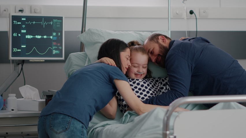 Happy parents visiting sick daughter hugging child during disease examination in hospital ward. Hospitalized kid wearing oxygen nasal tube resting in bed after suffering recovery surgery