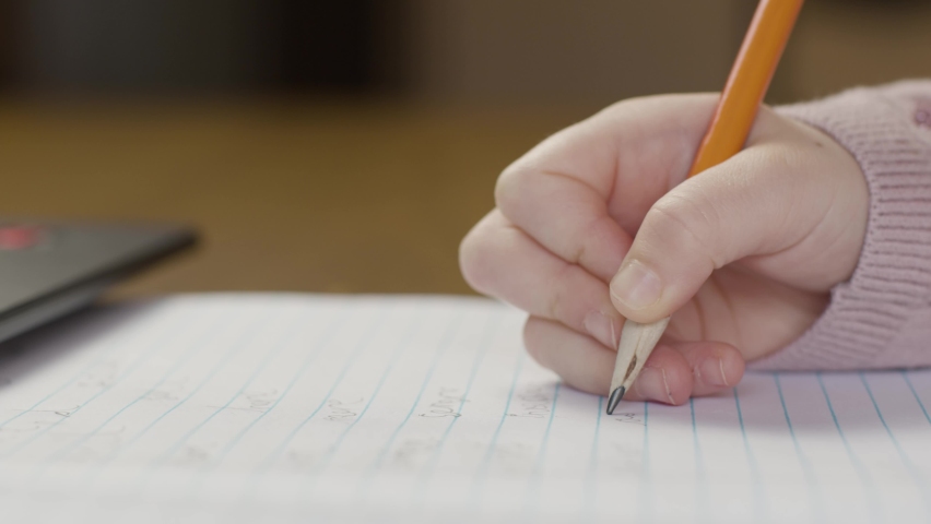 A close-up shot of a young child's hand writing on a piece of paper in front of a laptop. Royalty-Free Stock Footage #1075166477