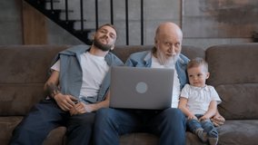 Happy family, father with elderly grandfather and cute child, sitting on couch with laptop and waving their hands in greeting gesture while looking at camera and smiling. Communication via video chat.
