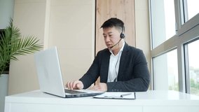 young Asian man with a headset conducts an online consultation, meeting or conference using a laptop and a webcam a video call. Male salesman in suit in office help center sale of goods Internet store