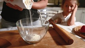 A woman pours white flour from paper packaging into a transparent glass bowl. Next to her sits her little daughter with a toy in her hands and watches the cooking process. Video materials