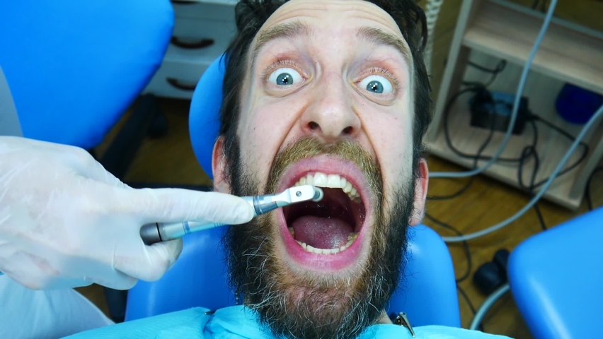 A young bearded man is afraid of professional brushing his teeth and screams in horror while sitting in the dentist's chair Royalty-Free Stock Footage #1075178438