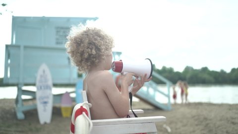 Young pretty curly boy sitting on high white chair with lifeline and posing with whistle on the beach against blue lifeguard tower.