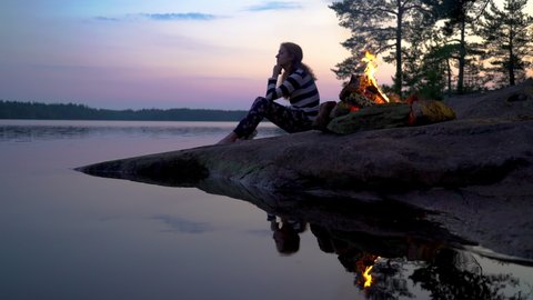 Young woman sitting dy the bonfire burning in a forest on the shore of a lake at midnight on the Summer Solstice. Summer White Night on the eve of Midsummer Day in Finland. Stockvideó