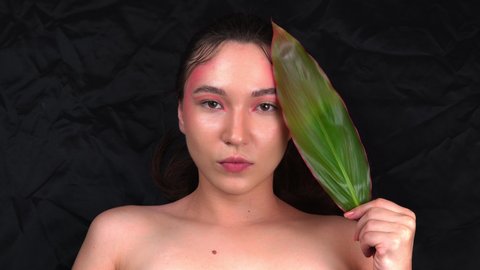 Beautiful young model posing for a photographer. A fashionable model with bright makeup holds a green leaf in her hands. Black fabric background. 4k footage