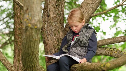 Tilt up of cute barefoot little girl sitting on tree branch in park or backyard and reading book aloud. Introvert kid spending summer time alone