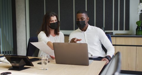 Diverse businessman and businesswoman wearing face masks discussing and using laptop in office. business and business people in office during covid 19 pandemic concept.