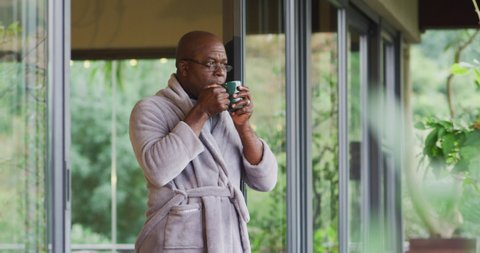 African american senior man standing on balcony wearing bathrobe drinking coffee and enjoying view. retirement lifestyle, spending time alone at home.