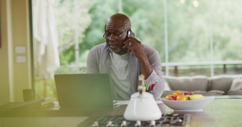 African american senior man in bathrobe sitting in kitchen using laptop and talking on smartphone. retirement lifestyle, spending time alone at home.