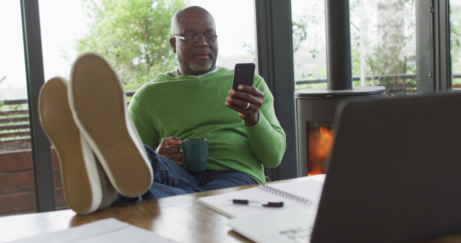 Smiling african american senior man relaxing with feet up, using smartphone and drinking coffee. retirement lifestyle, spending time alone at home.