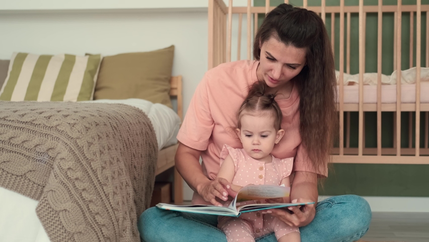 Childcare at Home, Child Protection, New Life, Leisure with Baby. Mom reads a book to the kid while sitting near the bed at home | Shutterstock HD Video #1075200728