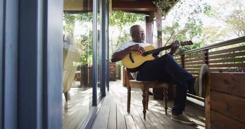 Thoughtful african american senior man sitting on balcony playing acoustic guitar. retirement lifestyle, spending time alone at home.