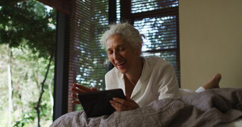 Senior mixed race woman lying in bed using tablet and smiling. retirement and senior lifestyle, spending time alone at home.