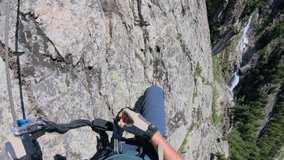 Male hiker ascends vertical via ferrata route. He clips cables and follows metal rungs while doing so