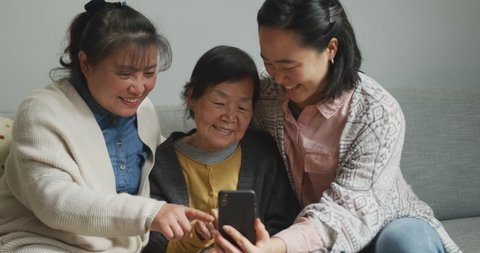 Smiling senior asian woman using smartphone with adult daughter and granddaughter embracing. multi generation family, senior lifestyle, togetherness and happiness concept.