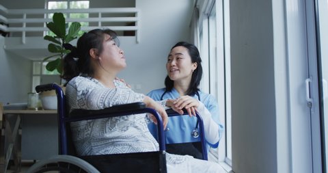 Smiling asian female doctor looking out of window and talking with female patient in wheelchair. medicine, health and healthcare services.