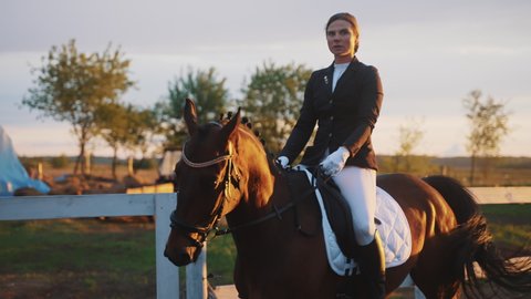 Competitive horsewoman riding on her dark bay horse. Girl with serious expression riding on her horse in the sandy parkour during the sunset time. The girl dressed in a black coat and white pants.