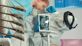 Vertical video: Dentist in protective equipment showing on tablet dental x-ray reviewing it with mother of kid patient. Medical team wearing face shield coverall, mask, gloves, explaining radiography