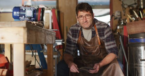 Caucasian male knife maker sitting in workshop, looking at camera and smiling. independent small business craftsman at work.