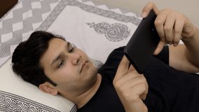 High angle shot - Young attractive teenager watching videos on his smartphone. Handsome Indian guy lying in his bed and using his mobile phone with a cute smile on his face - technology concept