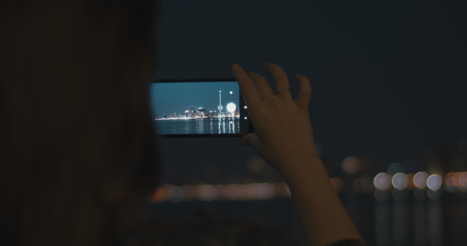 Close up of a young woman using a smartphone to take pictures of a night cityscape. Slow motion, rack focus, close up.