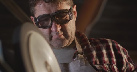Close up of caucasian male knife maker in workshop wearing glasses and using sander. independent small business craftsman at work.