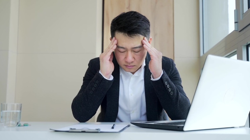 Young asian man, office worker sitting at workplace holding hands massages forehead and head with severe pain. Male at work with a headache. Up close face of sufferer is overloaded with sick. Closeup | Shutterstock HD Video #1075234976