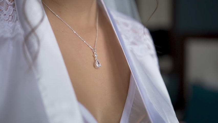 Morning of the bride. The girl stands in a white nightgown and satin robe and  with beautiful manicure on hand adjusts  jewelry hanging on a graceful neck