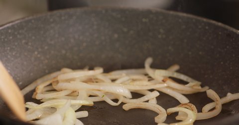 Stirring fried sliced onion in hot frying pan by wooden turner. Close up