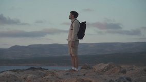 Young man with backpack takes photo on smartphone while walking along rocky coast near mediterranean sea on island of cyprus at sunset. Traveler with backpack takes pictures of beautiful sea views
