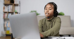 Funny asian girl talking with headset and learning with computer notebook in living room. Little girl studying online using her laptop at home. 