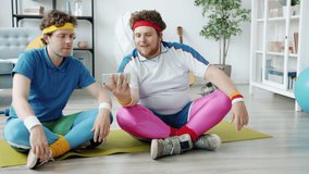 Slow motion of guys watching sports tutorial on smartphone screen and repeating movements sitting on yoga mat together wearing colorful sportswear.
