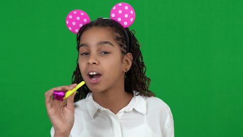 Excited happy cheerful African American teen girl wearing mouse toy ears blowing party whistle smiling on green screen. Portrait of joyful teenager celebrating birthday at chromakey background