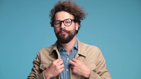 happy curly hair man in denim shirt with eyeglasses having fun, waving and laughing, fixing jacket and glasses, talking, nodding and flirting on blue background in studio