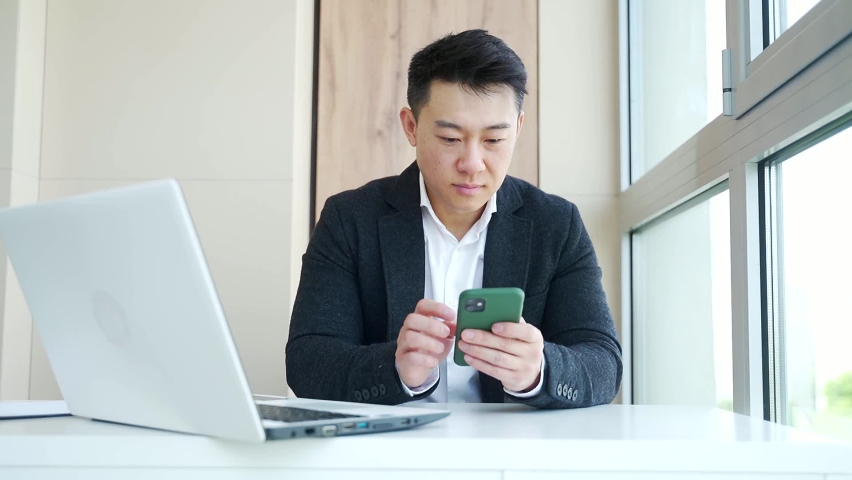Happy young Asian business man in office looking at mobile phone, with emotion winner or win, financial stock sports betting. Male joyfully exclaims playing game. Excited overjoyed celebrating success Royalty-Free Stock Footage #1075283786