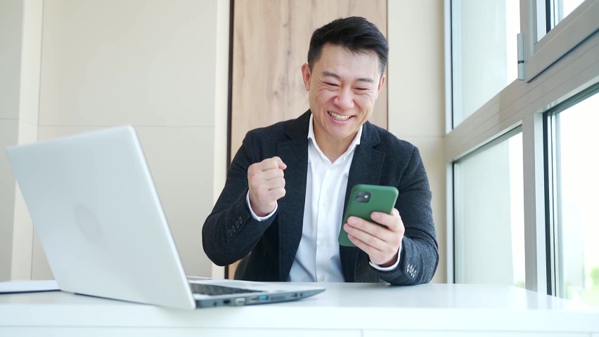 Happy young Asian business man in office looking at mobile phone, with emotion winner or win, financial stock sports betting. Male joyfully exclaims playing game. Excited overjoyed celebrating success | Shutterstock HD Video #1075283786