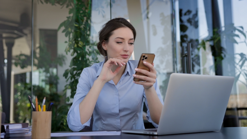 Young Girl Worker In The Office. She Is Sitting At The Table And Holding Mobile Phone In Her Hands. Girl Smiles.