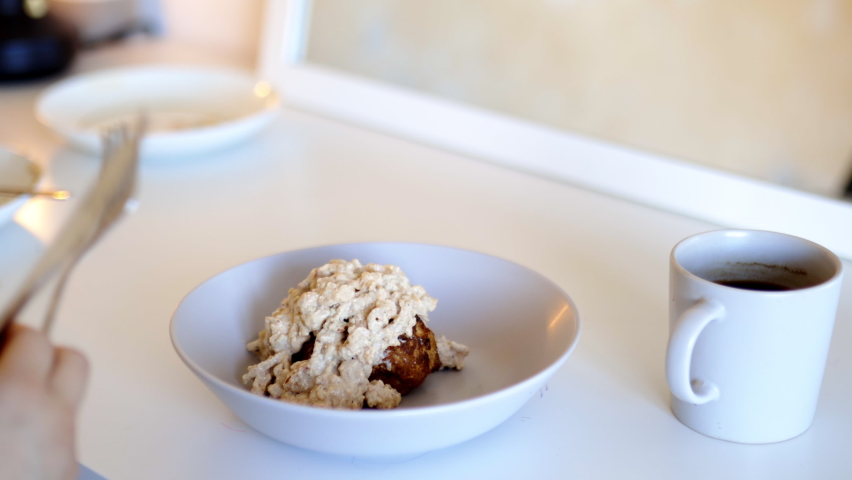 Female hands with fork and knife cutting biscuit covered in gravy in half. Woman sitting at table in front of a mirror ready to enjoy a meal. Traditional American breakfast